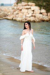 Beautiful young girl with long dark hair in a white long dress on the beach at sea in summer