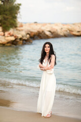 Beautiful young girl with long dark hair in a white long dress on the beach at sea in summer