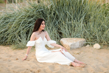 Beautiful young girl with long dark hair in a white long dress on the beach at sea in summer