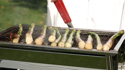 Fry asparagus on BBQ grill close up. Cooking fresh green asparagus or sparrow grass stems in outdoor wood logs charcoal grill with iron metal grate. Barbecued vegetables on fire for mixed vegan salad