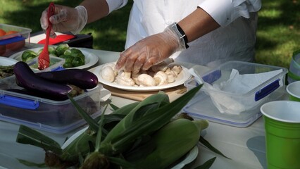 Woman chef in white short seasoning BBQ veggies for grill fry. Family adventures in the wild woods camping and picnic party. Preparation of food on the table for grilled mixed healthy salad.