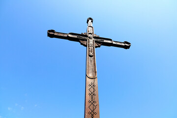 A wooden cross, intricately carved with a depiction of Jesus, stands against a clear blue sky. The...
