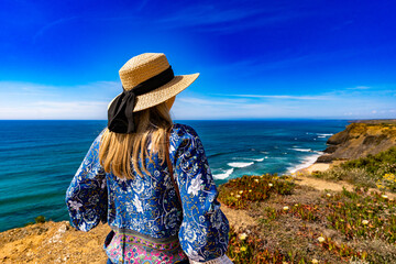 Mature woman tourist in sun hat standing on cliffs above ocean and enjoying views. Algarve Portugal. Back view