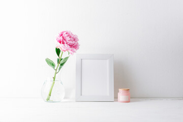 Minimal still life with pink peony in glass vase, blank picture frame and candle on white table against bright wall. Mockup