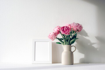 Pink peonies in ceramic jug and white frame on white table with shadow in natural light. Mockup