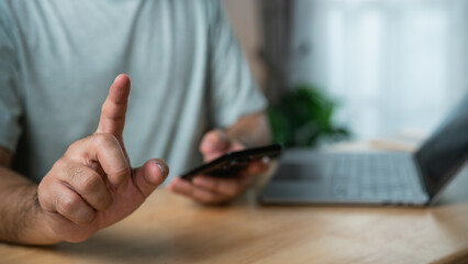 Person with mobile phone gesturing a stop signal while seated at a desk with a laptop and greenery in the background indoors