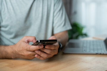 Person using smartphone while sitting at a wooden table next to a laptop with a serene indoor background in a cozy modern workspace