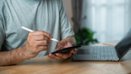 Man using stylus on smartphone while sitting at wooden desk beside laptop with plants in background, showcasing modern workspace and technology use