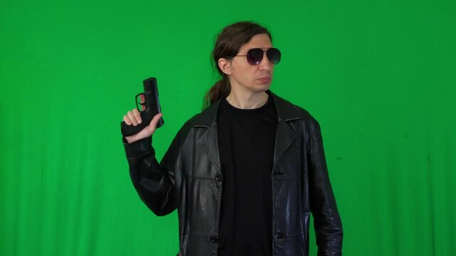 A mid-shot captures a serious and vigilant mood as a man in a leather jacket and sunglasses holds a handgun, looking to the side, ready for action, against a green screen