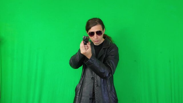 A mid-shot captures an intense and determined mood as a man in a leather jacket and sunglasses aims a handgun, focused on a target, against a green screen, ideal for action themes