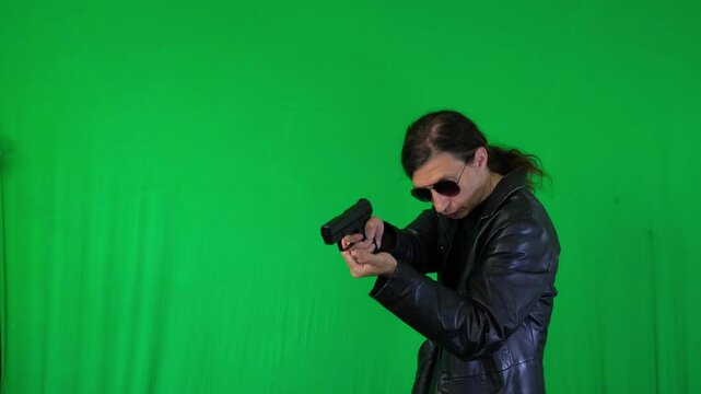 A mid-shot captures an intense and determined mood as a man in a leather jacket and sunglasses aims a handgun, focused on a target, against a green screen, ideal for action themes