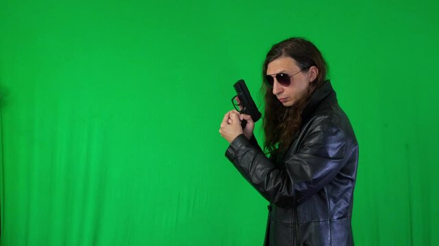 A mid-shot captures a serious and ready mood as a man in a leather jacket and sunglasses holds a handgun with both hands, looking directly at the camera, against a green screen