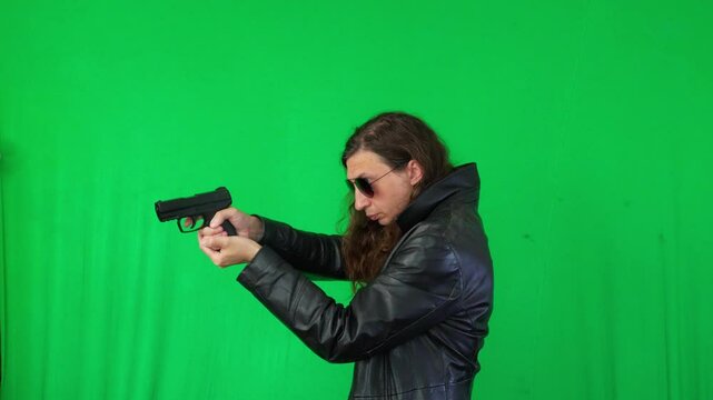 A mid-shot captures a serious and ready mood as a man in a leather jacket and sunglasses holds a handgun with both hands, looking directly at the camera, against a green screen