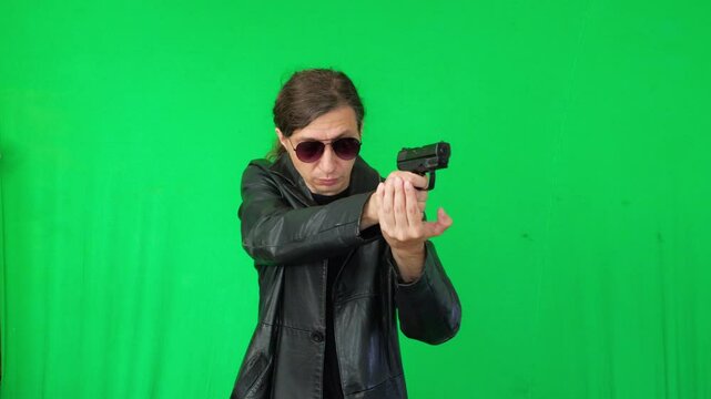 A mid-shot captures an intense and determined mood as a man in a leather jacket and sunglasses aims a handgun, focused on a target, against a green screen, ideal for action themes
