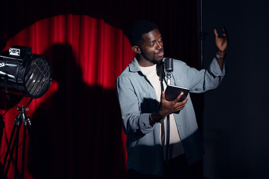 Happy young african american man comic tells stand up monologue on stage with red curtains. Concept humor standup comedian show open mike