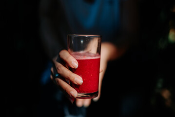 A hand holds up a glass of red juice in a dark and moody lighting setting, highlighting the drink.