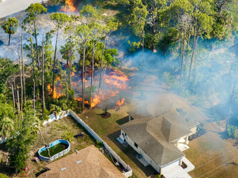 Flames from wildfire approach residential neighborhood in Florida with dense forest ablaze and smoke drifting over homes