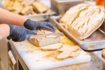 Baker slicing fresh artisan bread on a cutting board with a serrated knife. Close-up of hands in gloves, showing professional food preparation and hygiene standards.