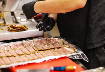 Worker in black gloves arranging and packaging sliced cured meat in a commercial kitchen. Food handling with hygiene focus during deli meat preparation.