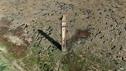 Historic brick tower rising from marshland with long shadow across cracked earth.