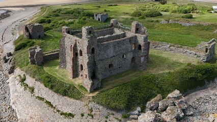 Drone view of crumbling coastal fortress surrounded by greenery and beach, captured in the UK.