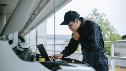 Mechanic inspecting car engine during cloudy day in parking garage while using laptop for...