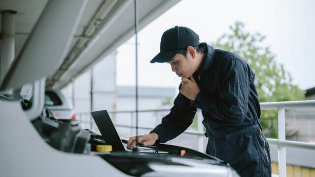 Mechanic inspecting car engine during cloudy day in parking garage while using laptop for diagnostics