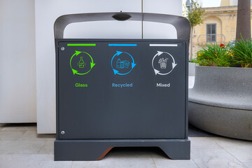 Modern public recycling bin with three labeled sections: glass (green), recyclable waste (blue),...