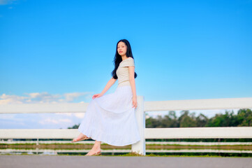 young woman in white skirt sitting on a white fence under bright blue sky, minimal countryside scene with peaceful and cheerful summer atmosphere.