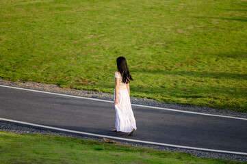 woman in a flowing white skirt walks alone on an empty road bordered by green grass, evoking a quiet sense of solitude and freedom
