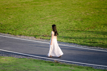 woman in a flowing white skirt walks alone on an empty road bordered by green grass, evoking a quiet sense of solitude and freedom
