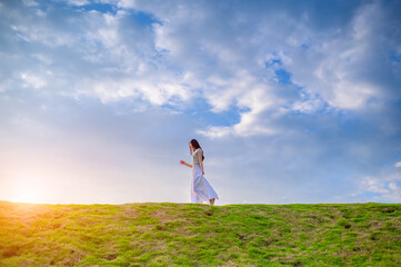 woman in white dress walking alone on green grassy hill under a bright cloudy sky at sunset, minimal composition with peaceful and serene natural atmosphere.