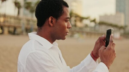 A young man in a white shirt takes photos or videos with his smartphone on the beach during a beautiful sunrise. The city is visible in the background. - Powered by Adobe