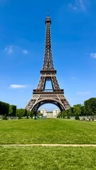 Fototapeta premium The full Eiffel Tower stands over a green lawn against a clear blue sky, with distant buildings visible through its arch and manicured trees flanking its base. 
