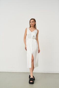 Elegant woman in fitted white dress with a front slit, posing confidently, wearing black platform sandals. She stands against a neutral backdrop, exuding sophistication and style