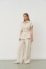 Fashionable woman poses in a stylish beige outfit featuring a belted top and wide-legged pants. She wears bold, round earrings and has a confident, poised expression