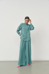 Woman in a teal button-up shirt and wide-leg pants, with hands in pockets. She wears pink shoes and...