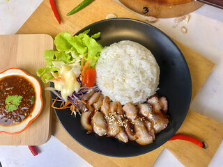 Appetizing Thai grilled pork neck with sesame seeds served with steamed jasmine rice and fresh salad including lettuce, purple cabbage, carrot, and cherry tomato. Presented on a black plate with spicy