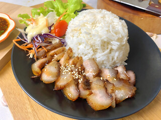 Appetizing Thai grilled pork neck with sesame seeds served with steamed jasmine rice and fresh salad including lettuce, purple cabbage, carrot, and cherry tomato. Presented on a black plate with spicy