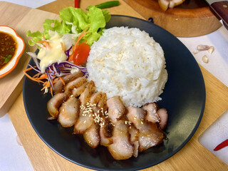 Appetizing Thai grilled pork neck with sesame seeds served with steamed jasmine rice and fresh salad including lettuce, purple cabbage, carrot, and cherry tomato. Presented on a black plate with spicy