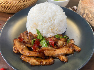 Delicious Thai grilled pork neck stir-fried with chili and holy basil served with steamed jasmine rice. Traditional Thai street food presented on a black plate with fresh red chili and garlic in the b