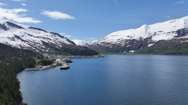 4k aerial video footage -flying across "Passage Canal", along Alaskan mountain Landscape in front of Whittier harbor and port area with Cityscape of Whittier and snow covered mountains in background. 
