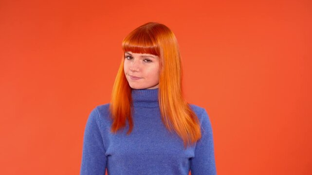 Young woman with vibrant red hair and a blue turtleneck sweater poses against a bright orange background, making a gesture with her arms crossed in an 'X' shape, conveying a message of refusal or disa