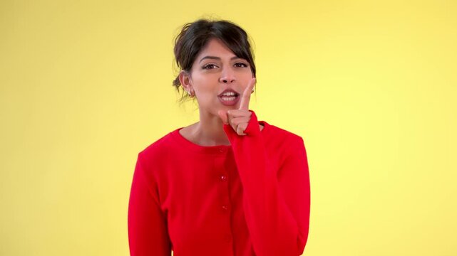 A woman in a red cardigan making a shushing gesture with her finger to her lips, set against a bright yellow background.
