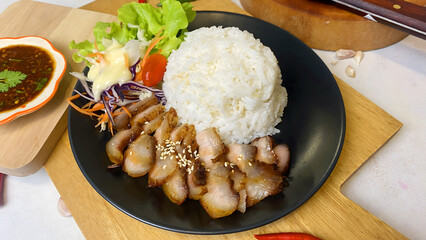 Appetizing Thai grilled pork neck with sesame seeds served with steamed jasmine rice and fresh salad including lettuce, purple cabbage, carrot, and cherry tomato. Presented on a black plate with spicy