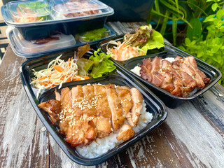 Grilled chicken served on steamed rice, garnished with sesame seeds, accompanied by fresh lettuce, shredded cabbage, and carrot salad with mayonnaise dressing. Presented in a black takeaway box on a r