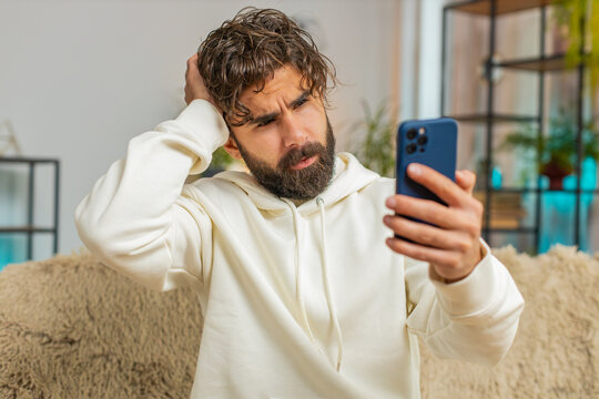 Upset displeased Indian man using smartphone sitting on sofa couch in living room. Young adult Hispanic guy in hoodie touching head loses becoming surprised by sudden lottery results or bad news.