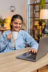 Plakat Young Indian woman working on laptop computer drinking cup of warm hot coffee sits at table home office room in the morning. Hispanic female freelancer enjoying relaxing, taking a break. Vertical.