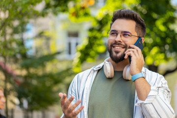 Caucasian young male man in glasses tourist talking cellphone smiling laughing. Guy in casual using mobile phone call happy discuss news friend smartphone communication on city town street outside.