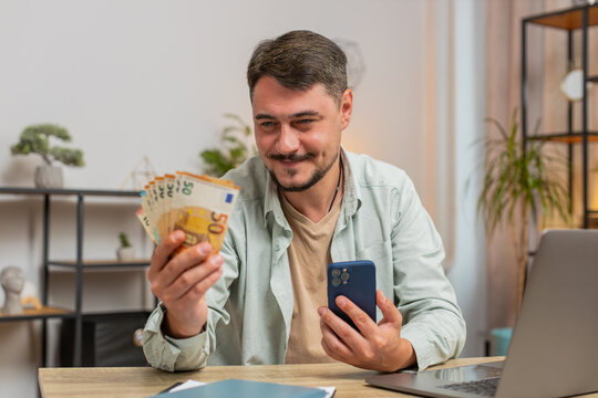 Planning family budget. Smiling Caucasian man counting money euro cash use smartphone calculate domestic bills. Guy satisfied of income, saves salary money at home office table workplace. Lifestyle
